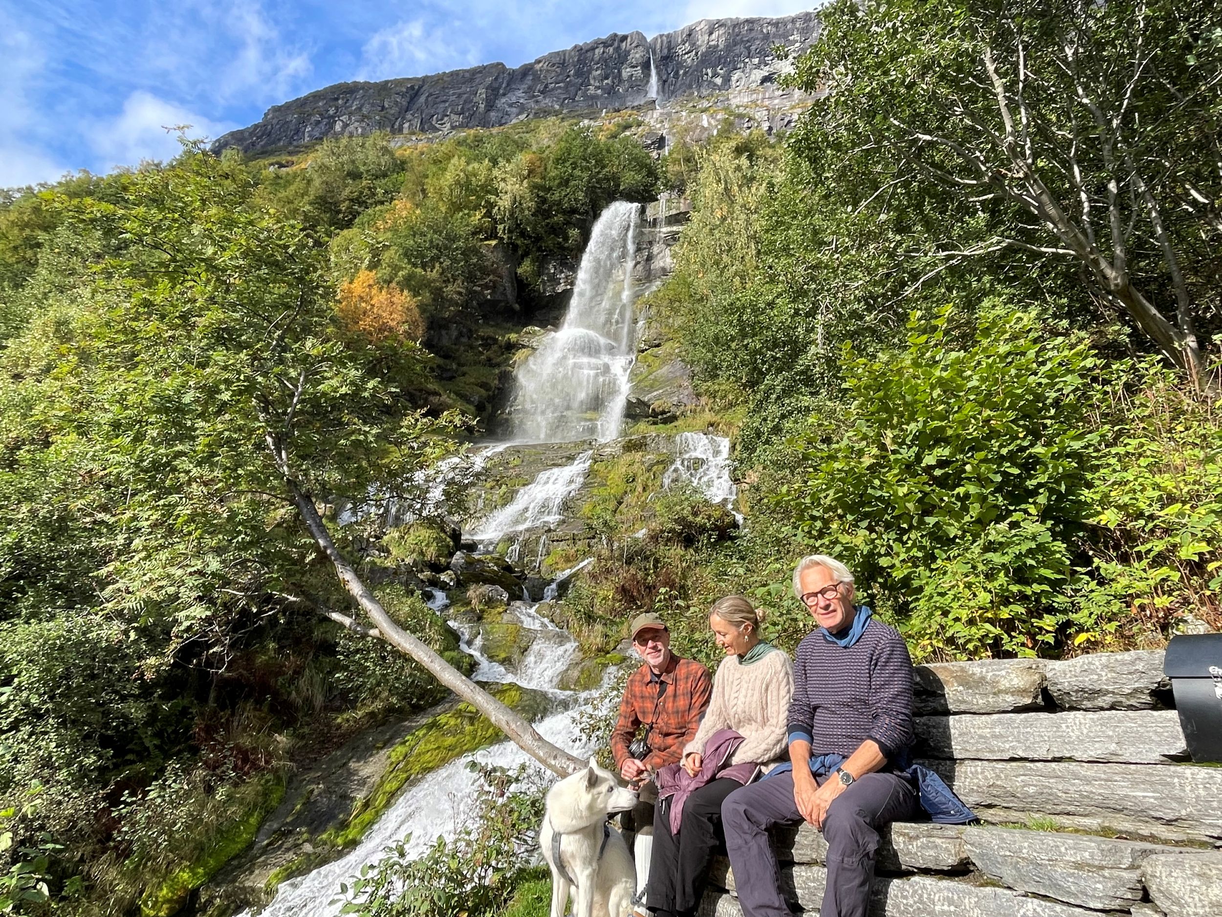 Tre personer og en hund på steinbenk foran høyt fossefall