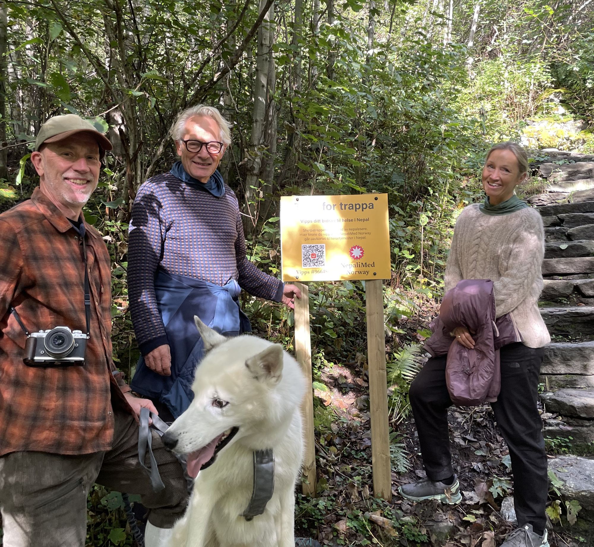 Tre personer og en hund foran gult skilt i naturen