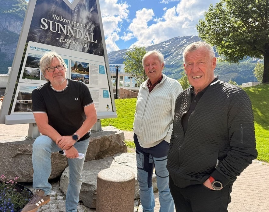 Tre menn foran et skilt med kulturhus, fjell og blå himmel bak.